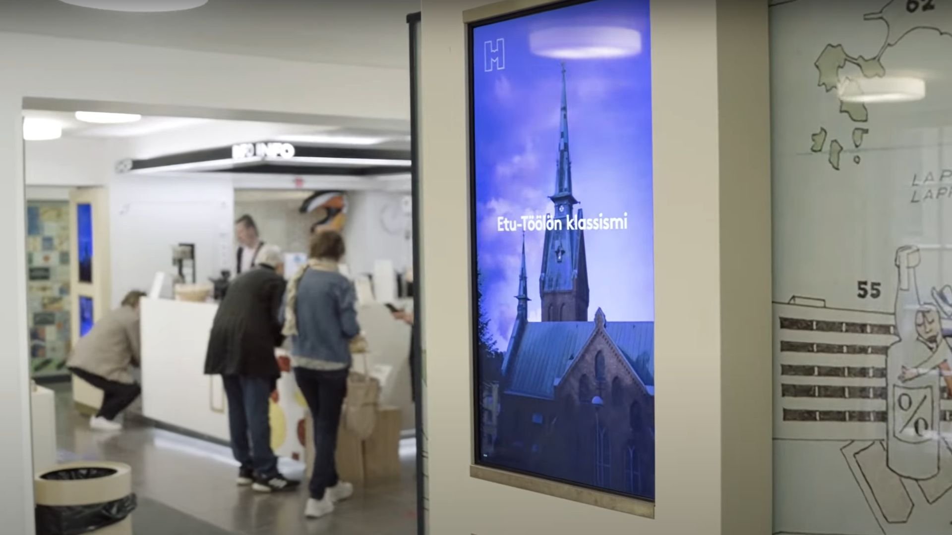 Helsingin Oodin infonäyttö. Kuvan taustalla henkilöitä palveltavina infopisteessä.
